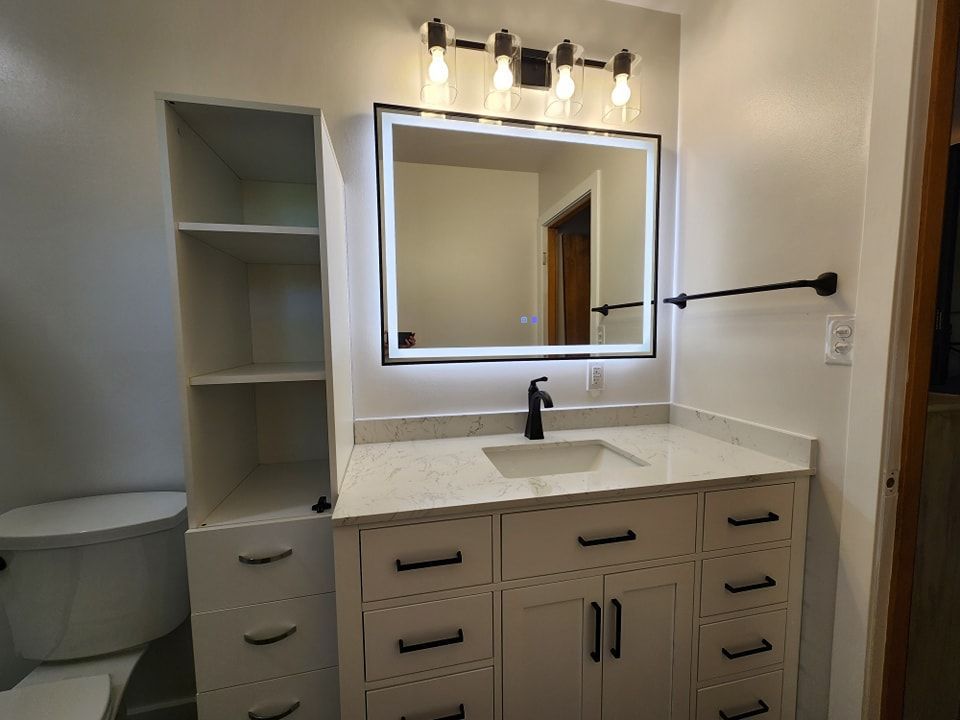 Modern bathroom with white vanity, black hardware, lit mirror, and open storage.