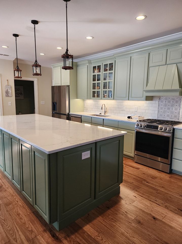Green and white kitchen with island, pendant lights, stainless appliances, and hardwood floors.