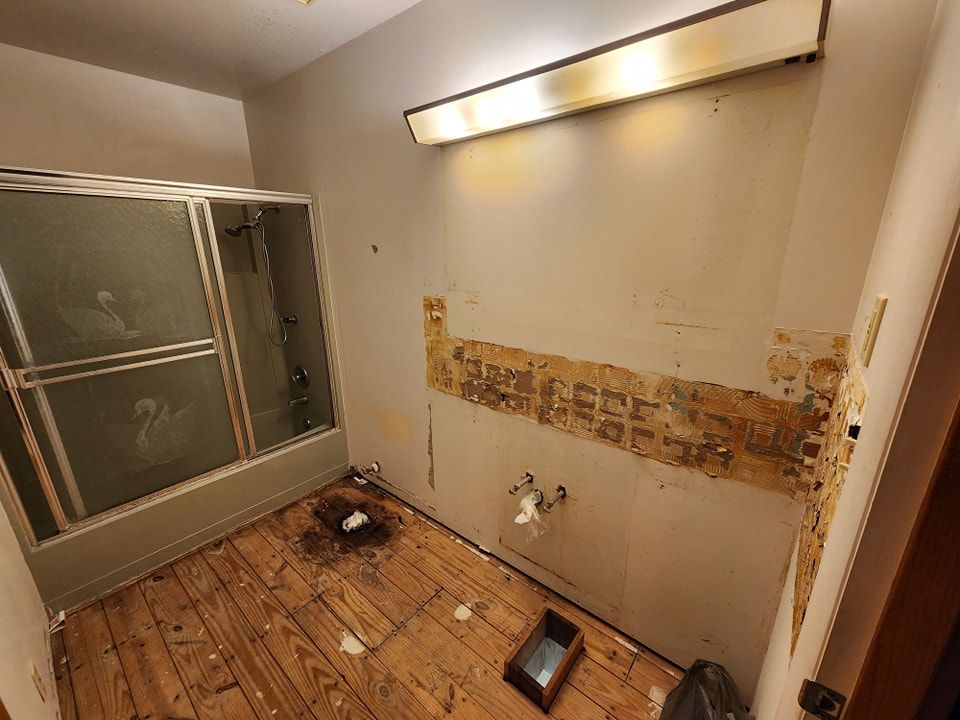 Bathroom renovation in progress, damaged wall, exposed wood, shower stall, dirty floor, overhead light.