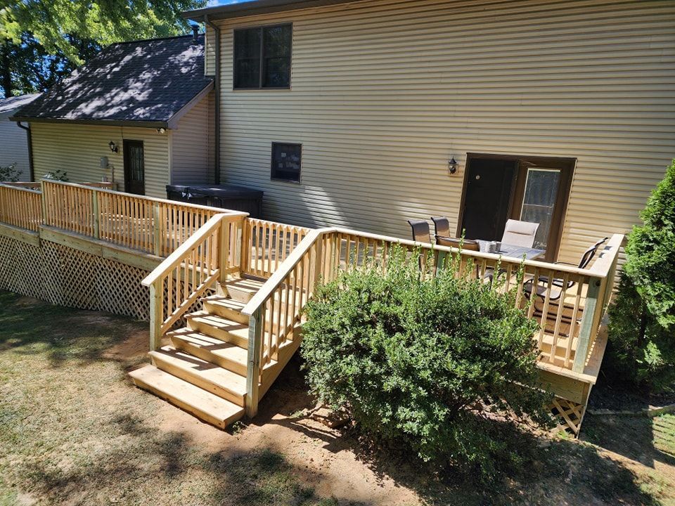 Wooden deck with stairs attached to a beige house, next to greenery on a sunny day.