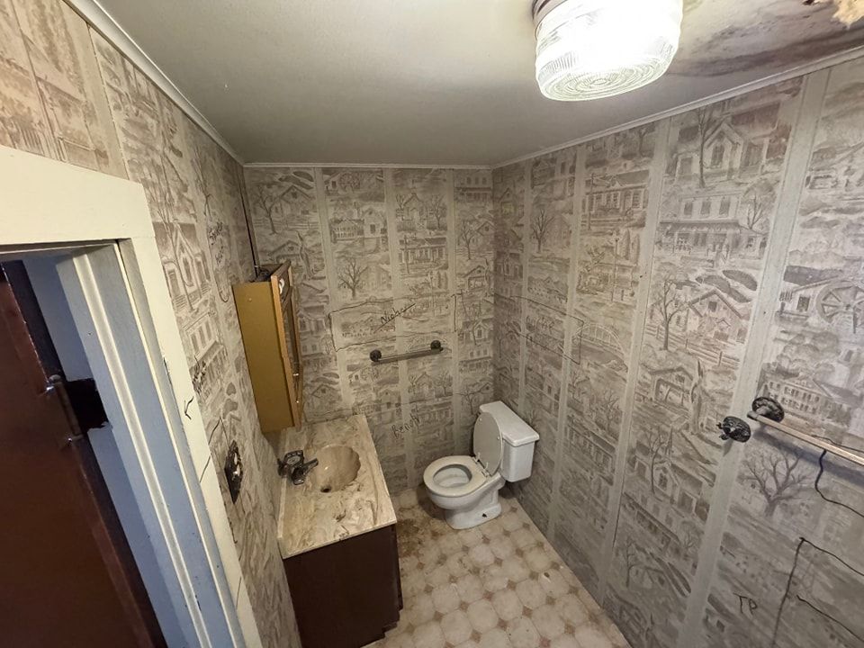 Small bathroom with patterned wallpaper, toilet, sink, and cabinets. A doorway is visible.