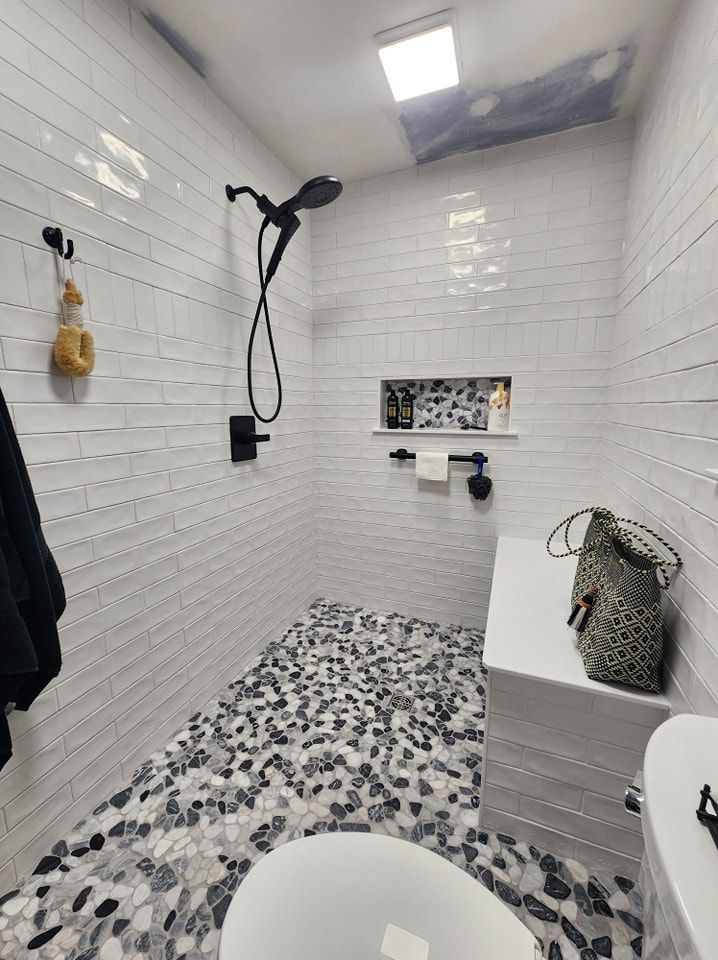 Modern shower with pebble floor, white tile walls, black fixtures, and a bench.