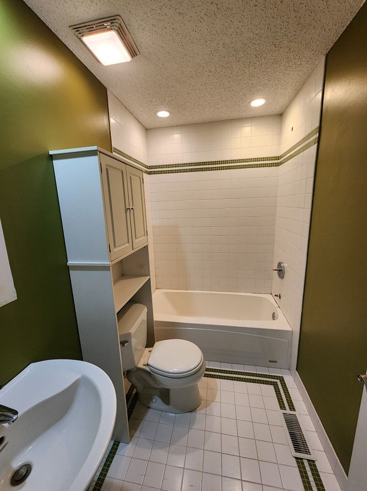 Bathroom with white tile tub surround, green walls, white toilet and sink, and white cabinet.
