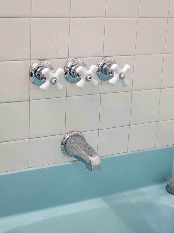 White-tiled bathroom wall with three white-handled faucets above a chrome spout and turquoise tub.