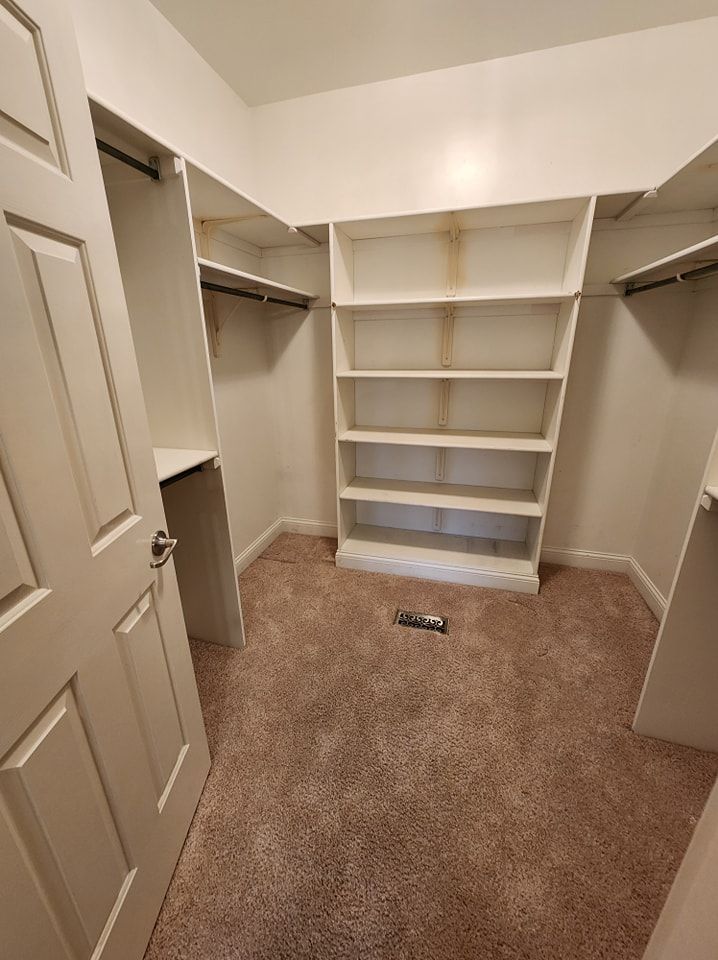 Empty walk-in closet with shelves and hanging rods. Beige walls, brown carpet, and white built-ins. Door on the left.