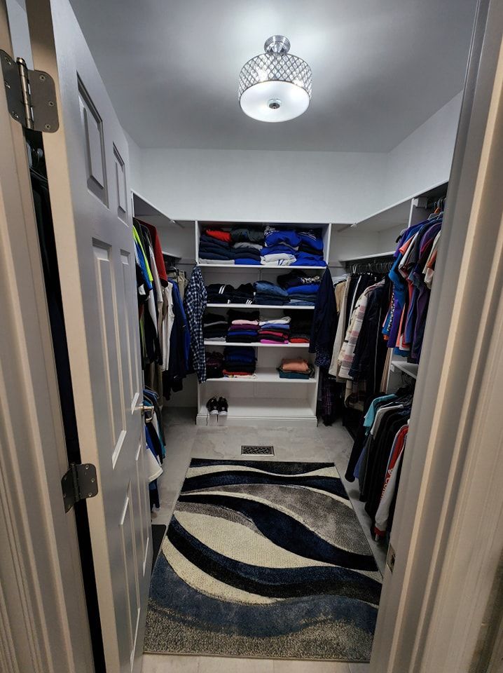 Walk-in closet with clothes on racks and shelves, blue and gray rug, overhead light, and white walls.