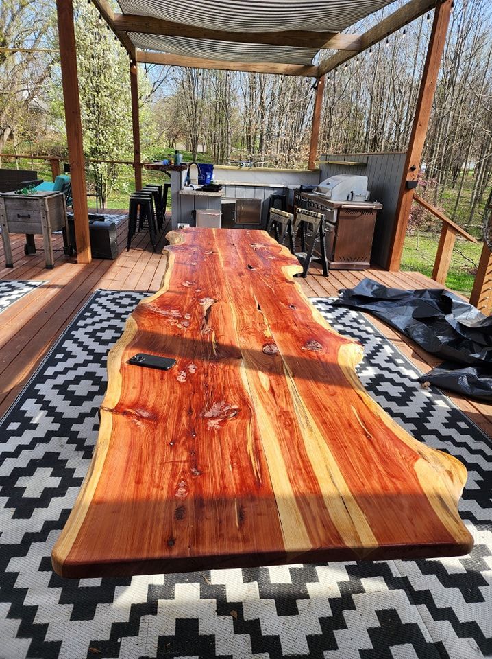 Large wooden live-edge table on an outdoor deck with a black and white rug, surrounded by a pergola and kitchen area.