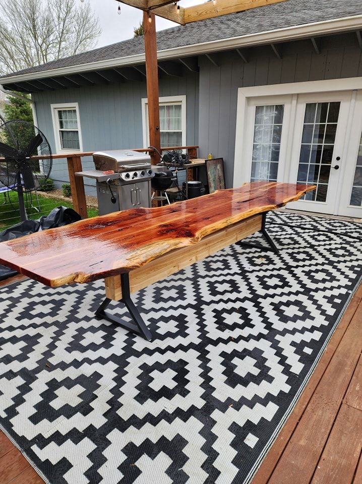 Live-edge wooden table on deck, with a grill and house in the background. Black and white rug.