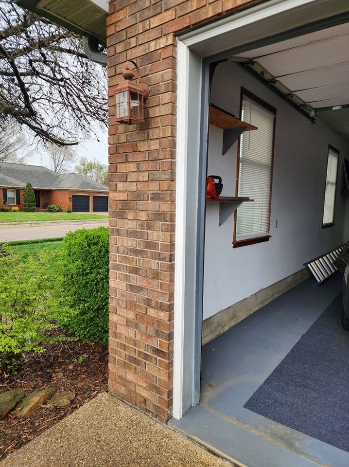 Garage opening. Brick pillar with a light fixture. Interior shows shelves, window, and rug.