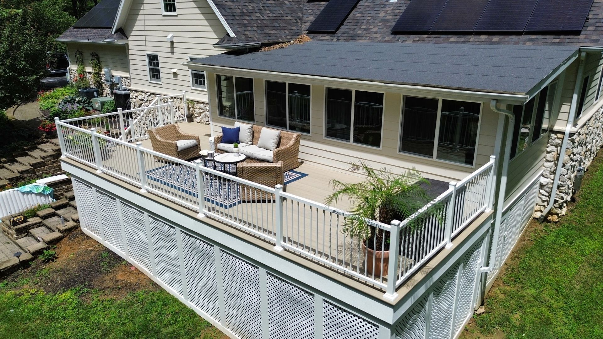 A deck with white railings and lattice, furnished with seating and a rug, attached to a house with a sunroom.