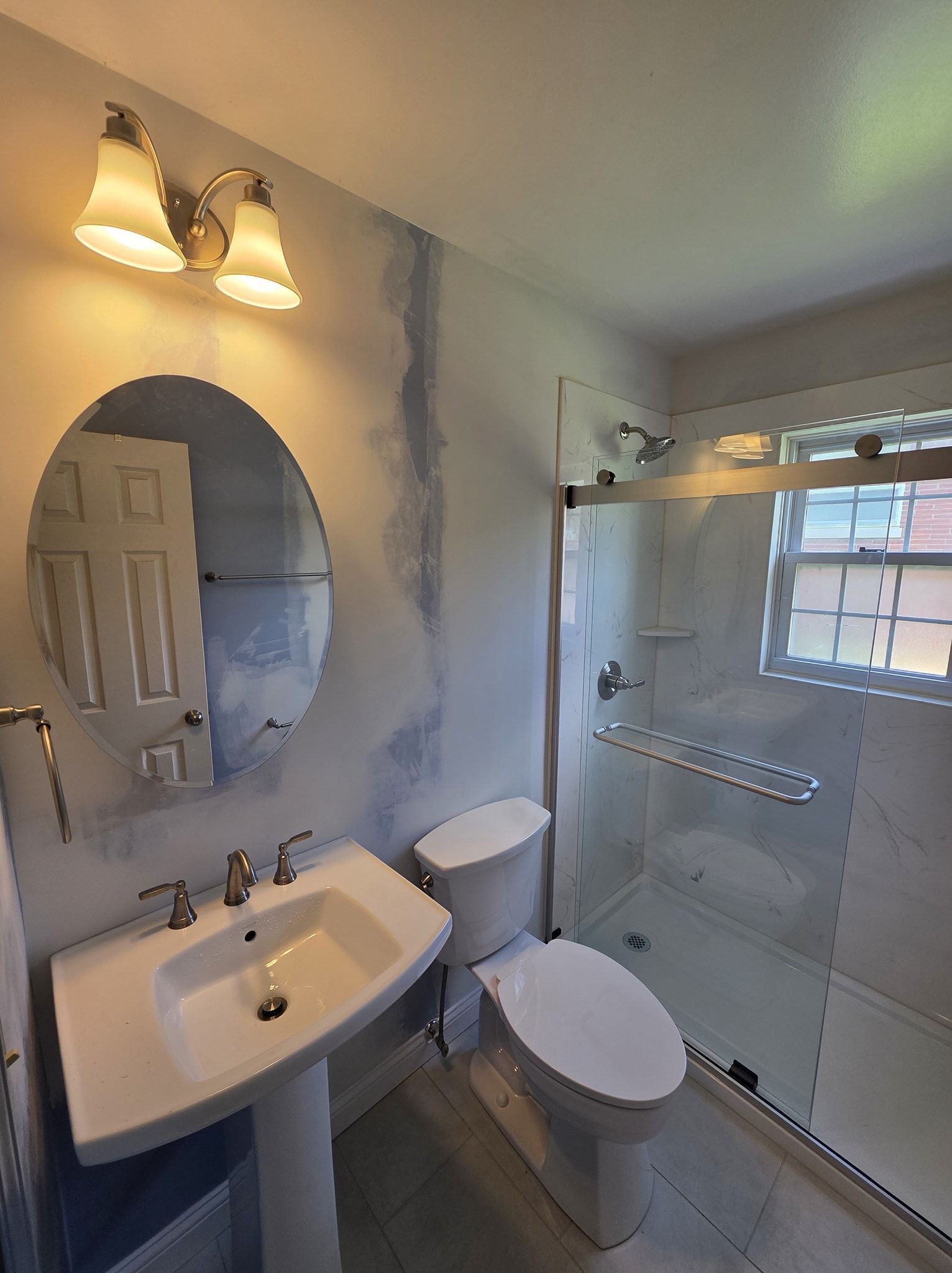 Bathroom with white pedestal sink, toilet, and glass shower. Blue and white walls, oval mirror, and three-light fixture.