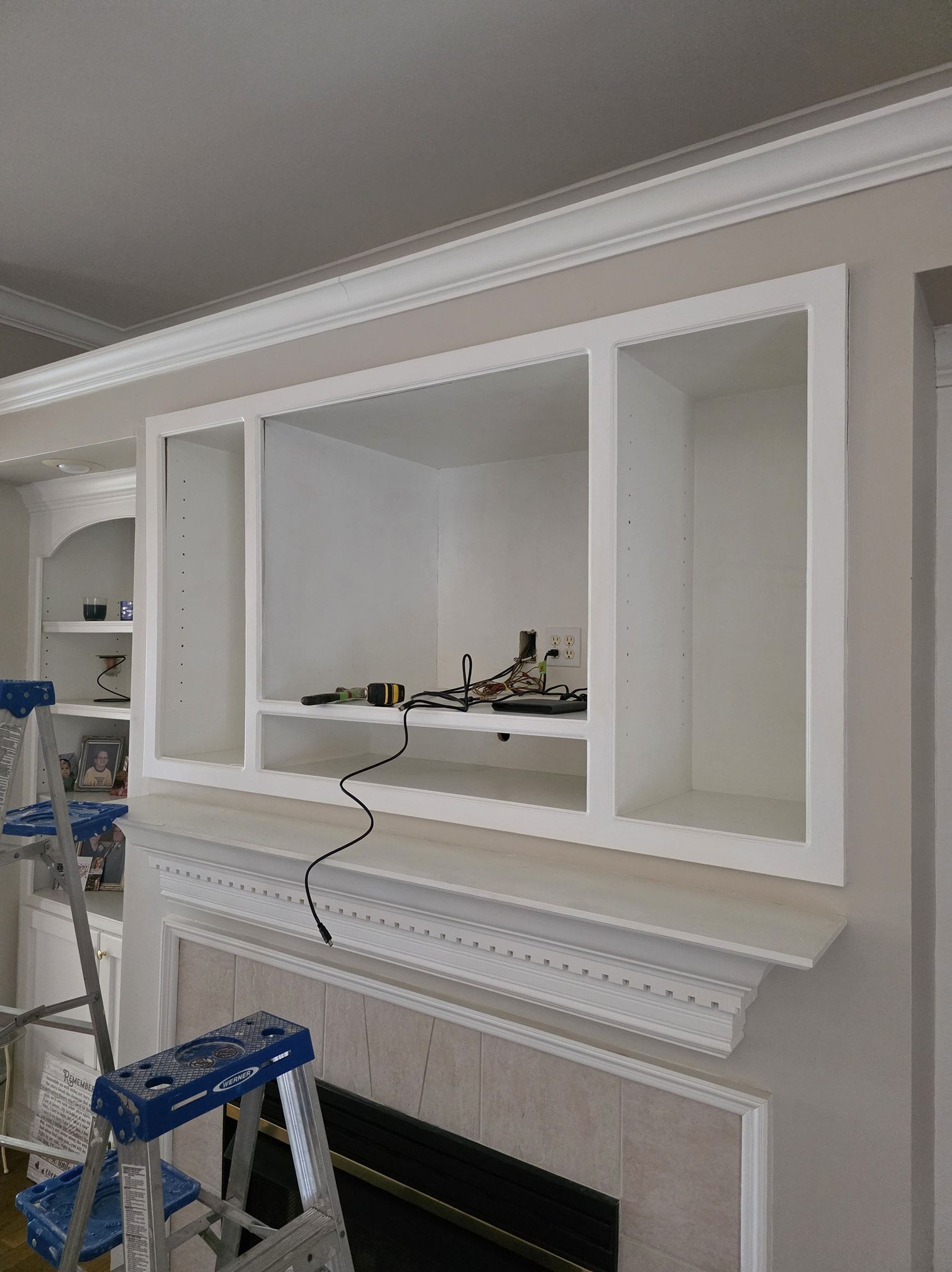 White built-in cabinet above a fireplace. Wires visible inside the cabinet. A step ladder is in the foreground.