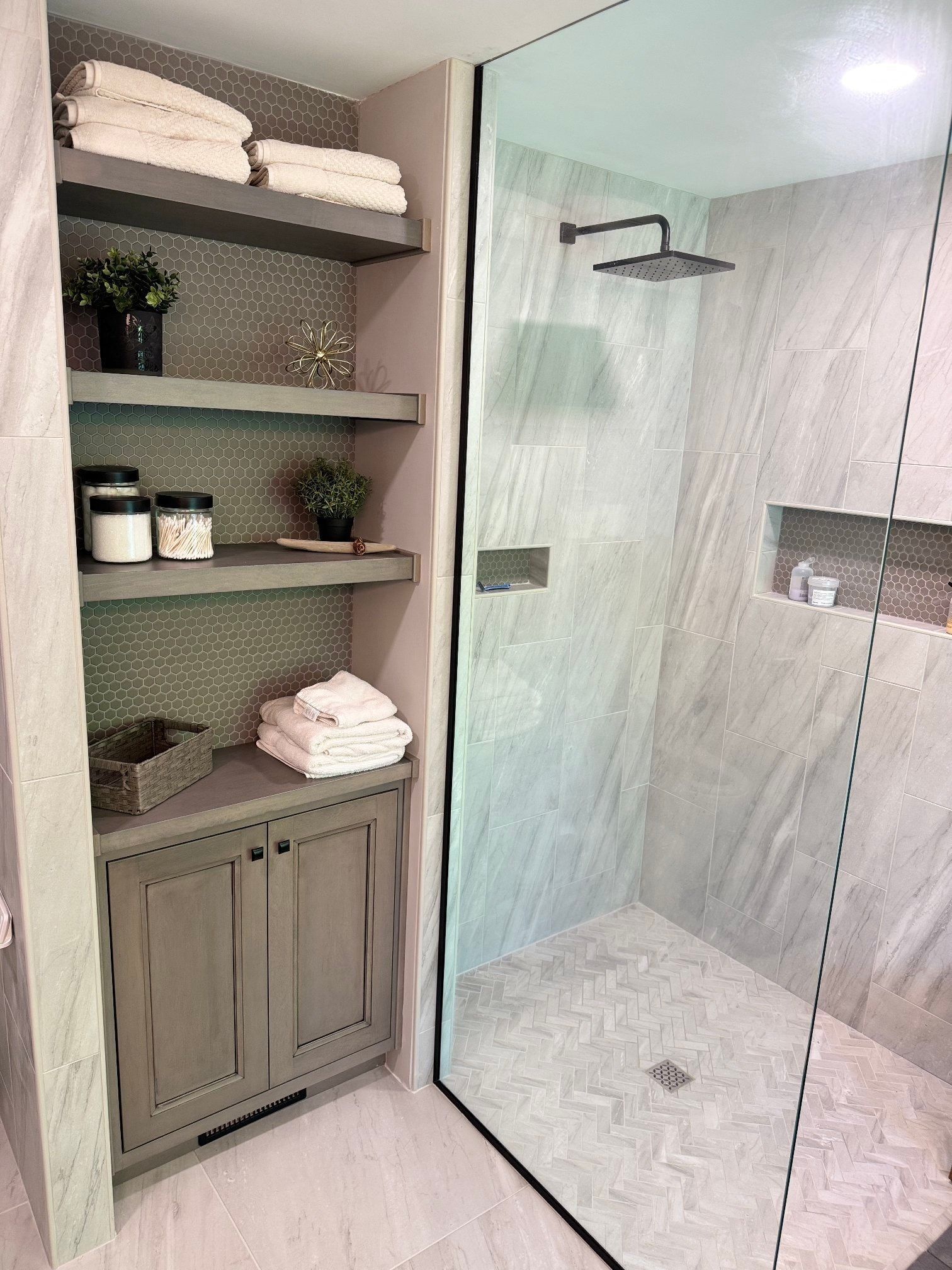 Bathroom with glass shower, built-in shelves holding towels, toiletries, and plants. Gray, white tones.