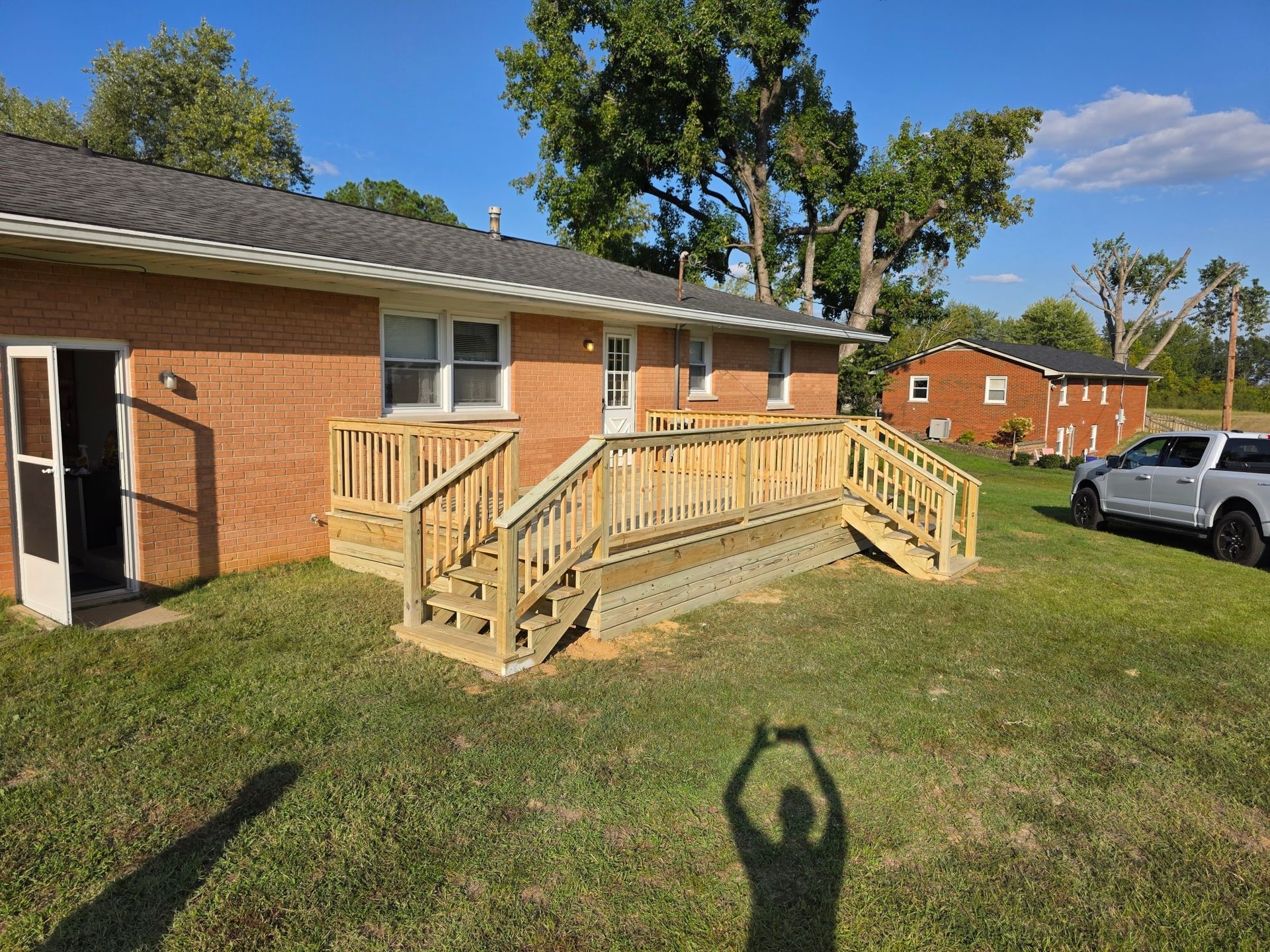 Brick house with wooden deck and ramp; person's shadow in foreground, truck in the distance.