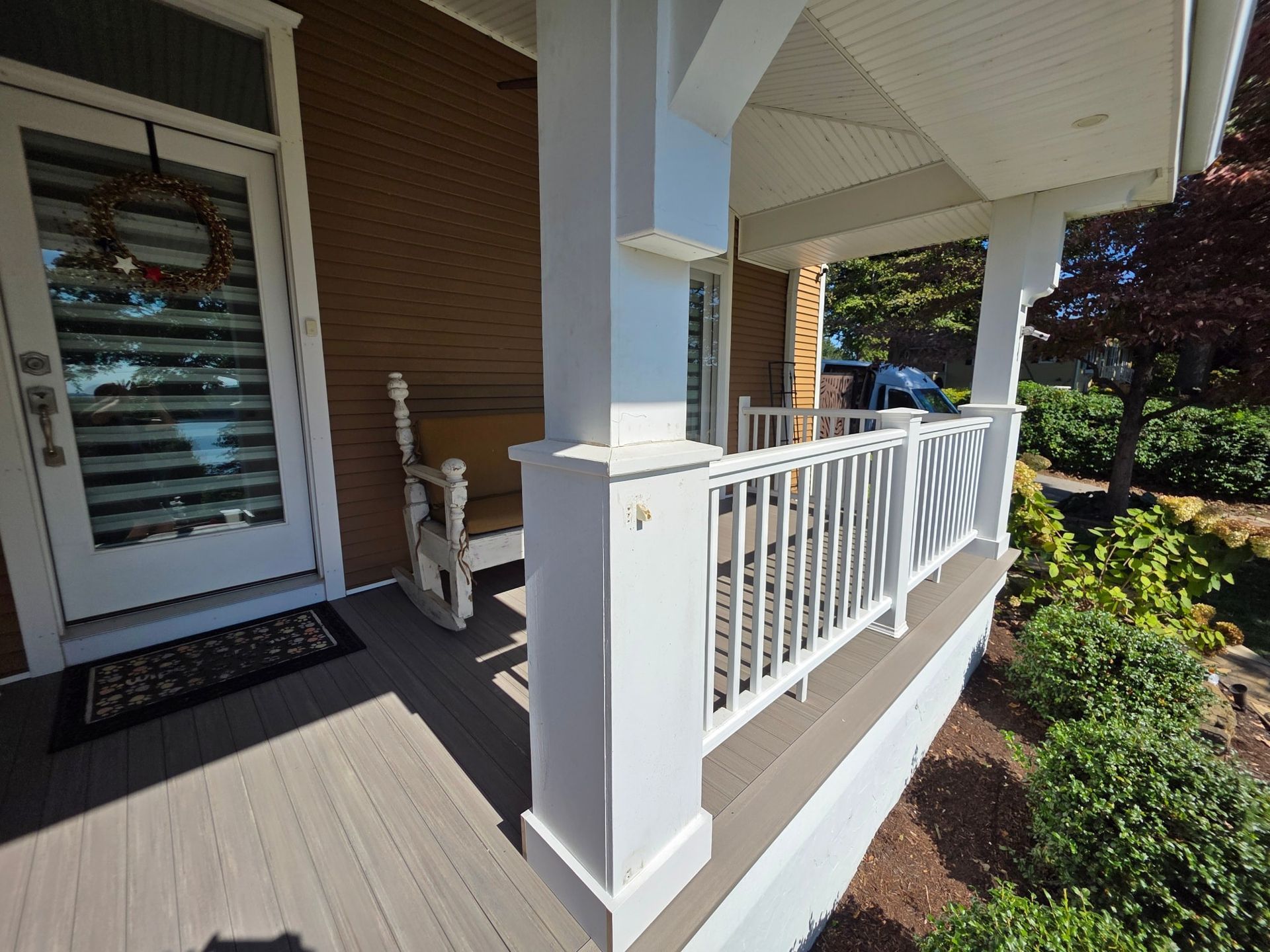 White porch with columns, railing, and front door; rocking chair on the porch, bushes and a tree in the yard.