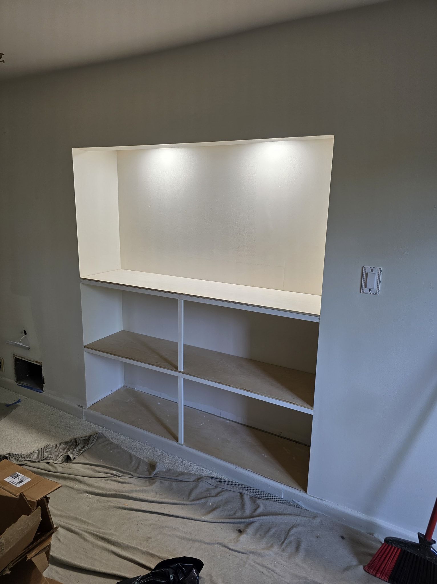 White built-in shelves in a recessed wall, with two lights overhead.  Paint and a drop cloth on floor.