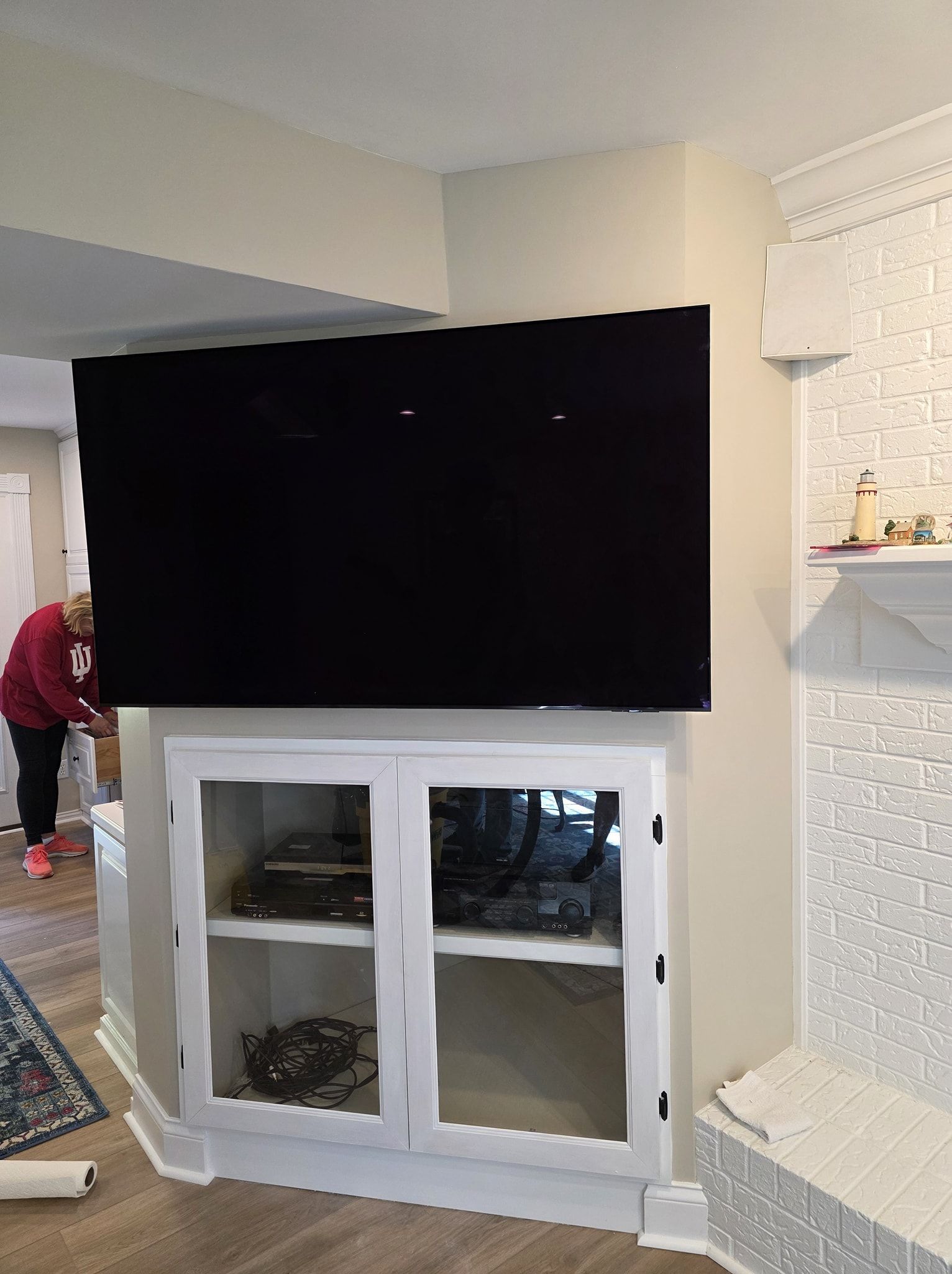Black TV mounted above a white cabinet. Person in the background. Corner of a room.