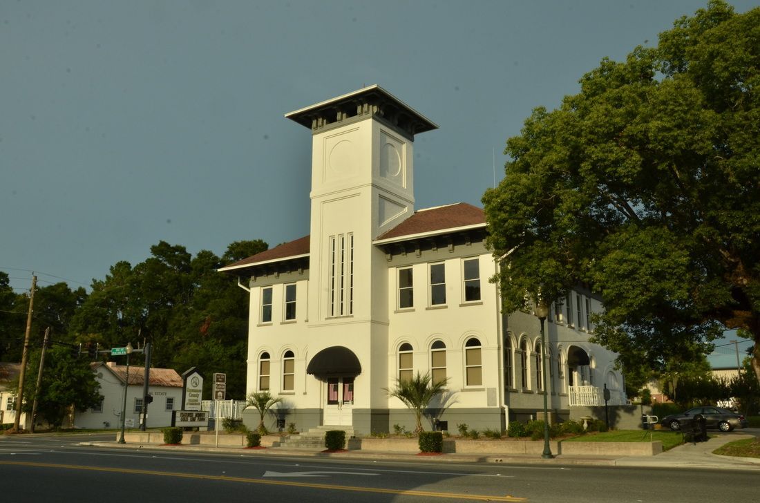White, two-story building with a tower, a brown roof, and arched windows. Street view with trees and a cloudy sky.