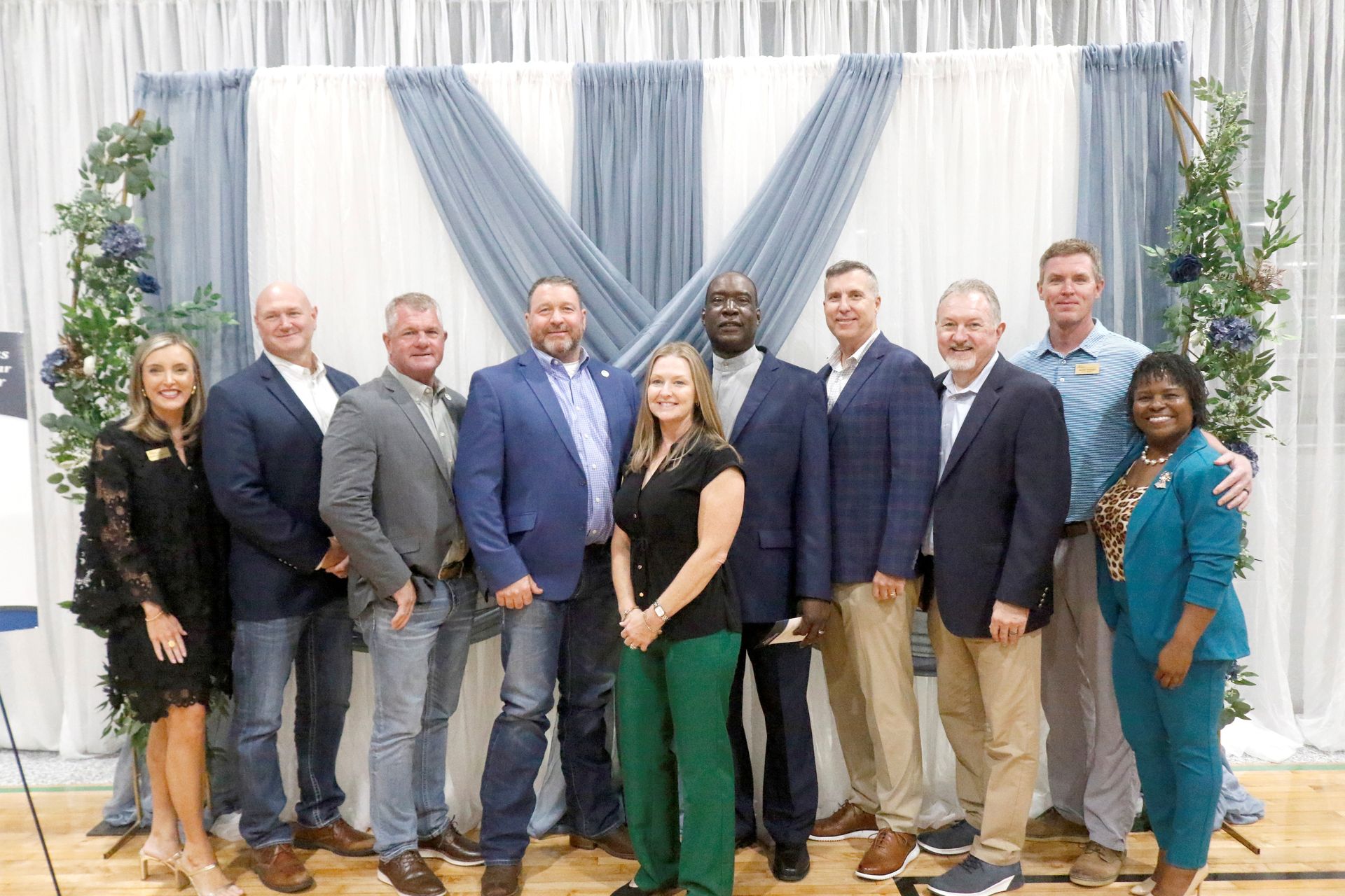 Group of men in business attire, standing indoors in front of a backdrop, smiling.
