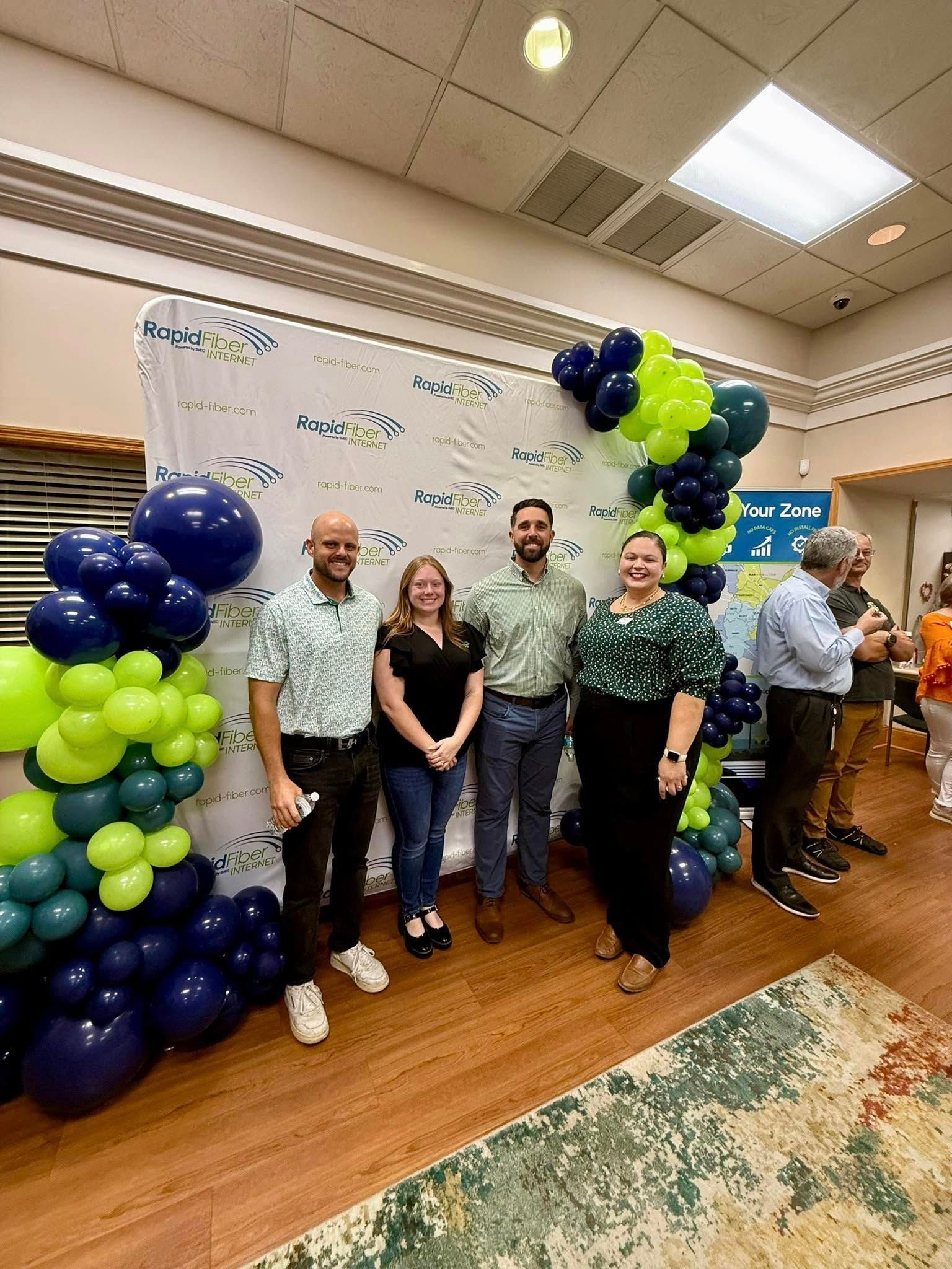 Group cuts ribbon at an event hosted by The Chamber. Brick building, pink ribbon, many people smiling.