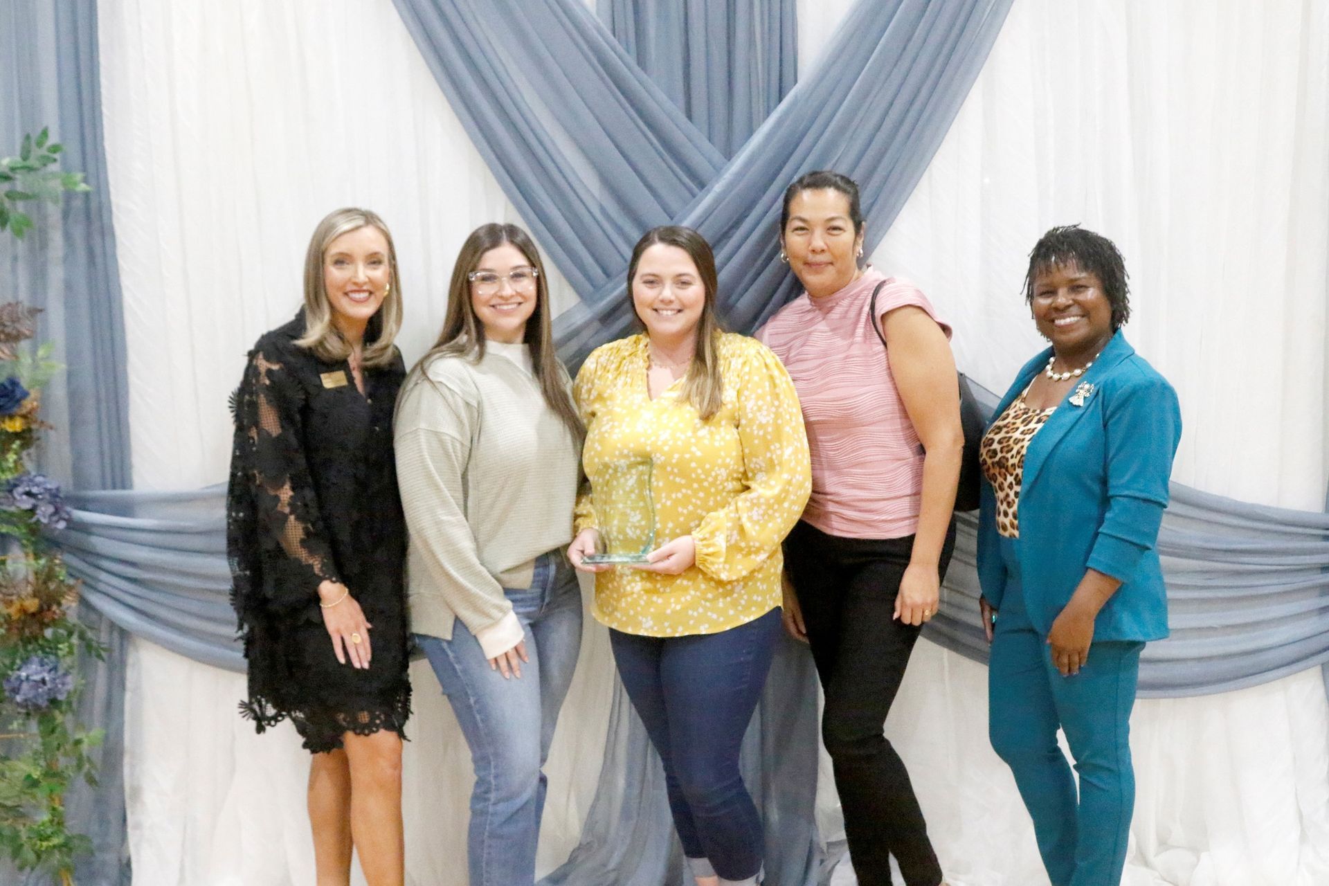 Group of people smiling, holding a blue award. Indoor setting with a backdrop, logo, and a blue and white theme.