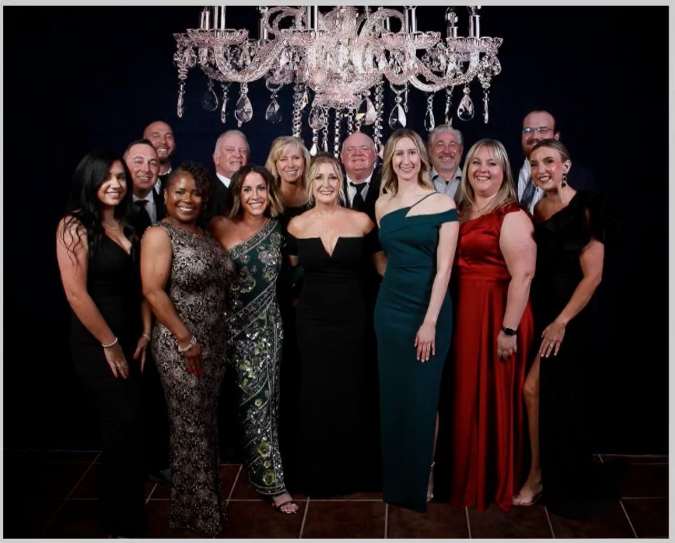 Group of people posing for a photo under a chandelier; formal attire; dark background.