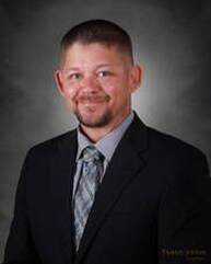 Man in a dark suit, light blue shirt, and patterned tie, smiling against a gray backdrop.