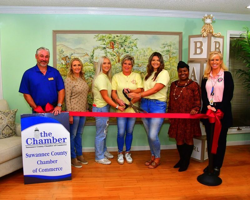 Ribbon-cutting ceremony at a business; people in a room, cutting a red ribbon, Suwannee County Chamber of Commerce.