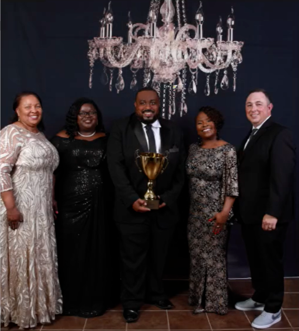 Group of five people posing, one holding a trophy, in formal attire, under a chandelier.