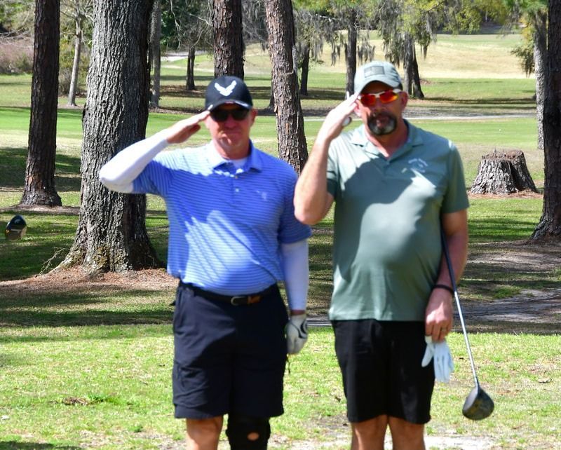 Two men on a golf course saluting. The man on the left wears blue and black. The man on the right wears green and black.