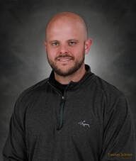 Bald man with a trimmed beard smiles, wearing a gray zip-up sweatshirt, against a blurred gray background.
