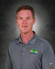 Man in gray polo shirt with logo, smiling, against a blurred gray backdrop.