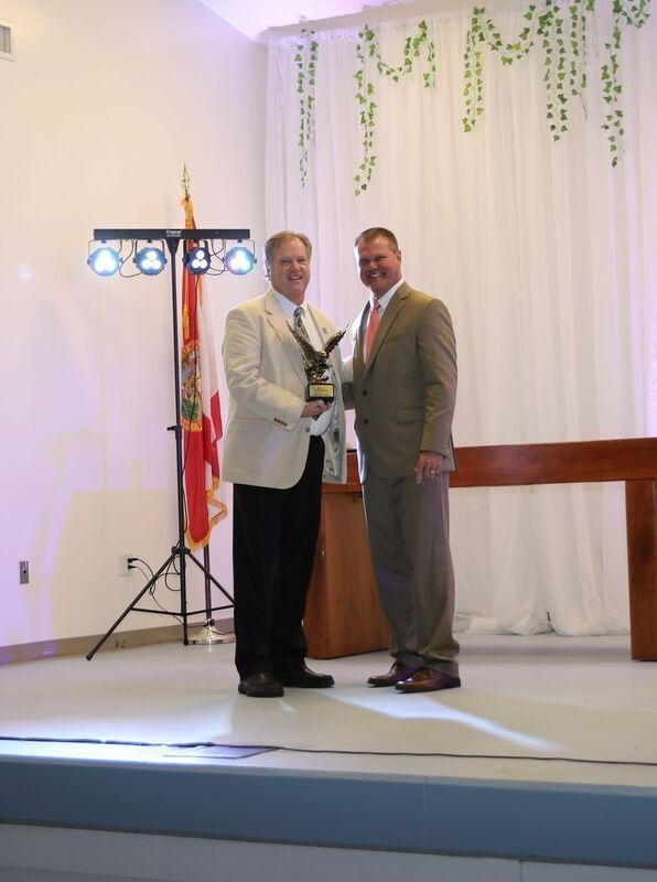 Two men on stage holding award; Florida flag and lights in the background.