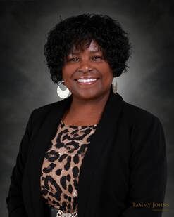 Woman with curly black hair, wearing a black blazer, animal print top, and silver earrings, smiles at the camera.