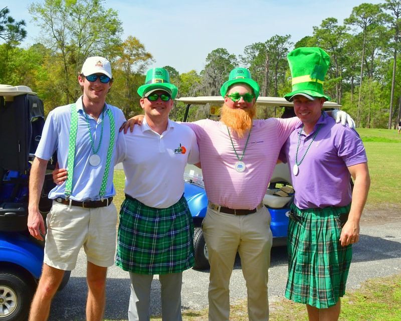 Four men in St. Patrick's Day attire posing with golf carts; green hats, kilts, beards, and sunglasses on a golf course.