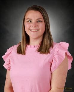Woman with light skin wearing a pink ruffled top smiling at the camera, against a dark background.