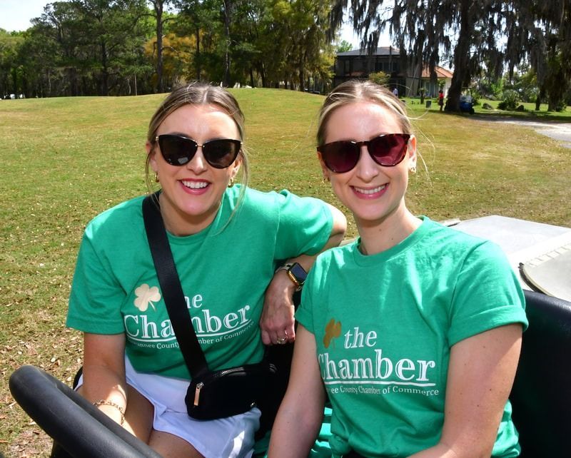 Two women in green t-shirts and sunglasses smile from a golf cart.