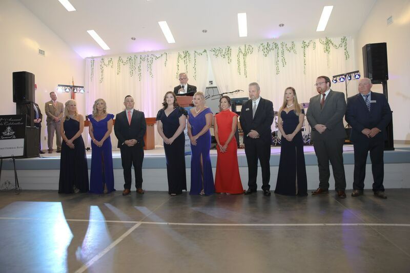 Group of people in formal attire on a stage, some are holding microphones, with a decorative backdrop.