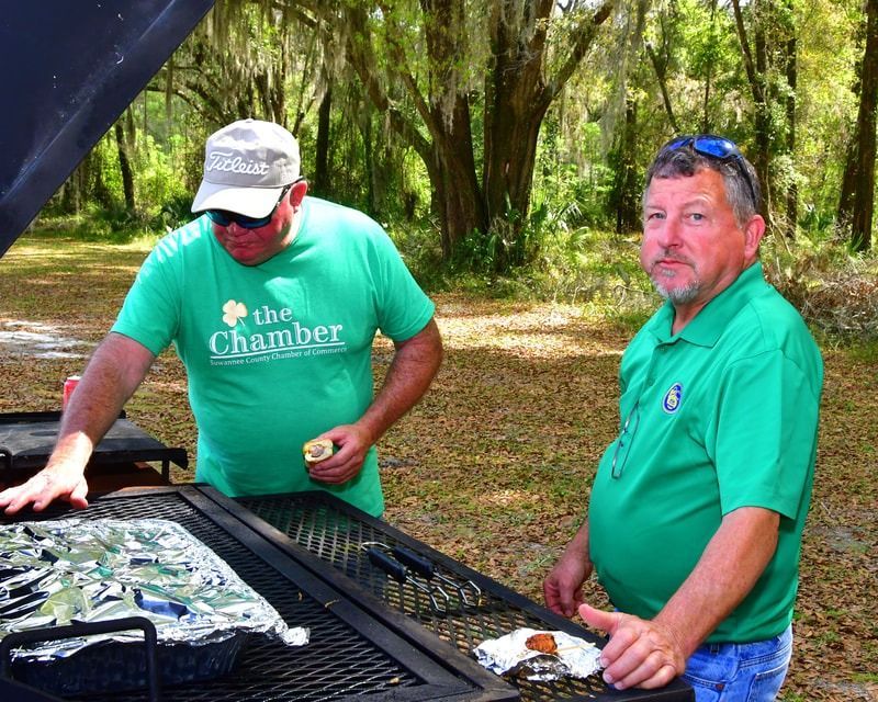 Two men grilling outdoors. One in a green shirt is holding food, the other is next to the grill.