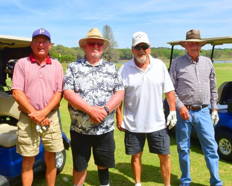 Four men pose in front of golf carts on a course; two wear hats, one holds a club.