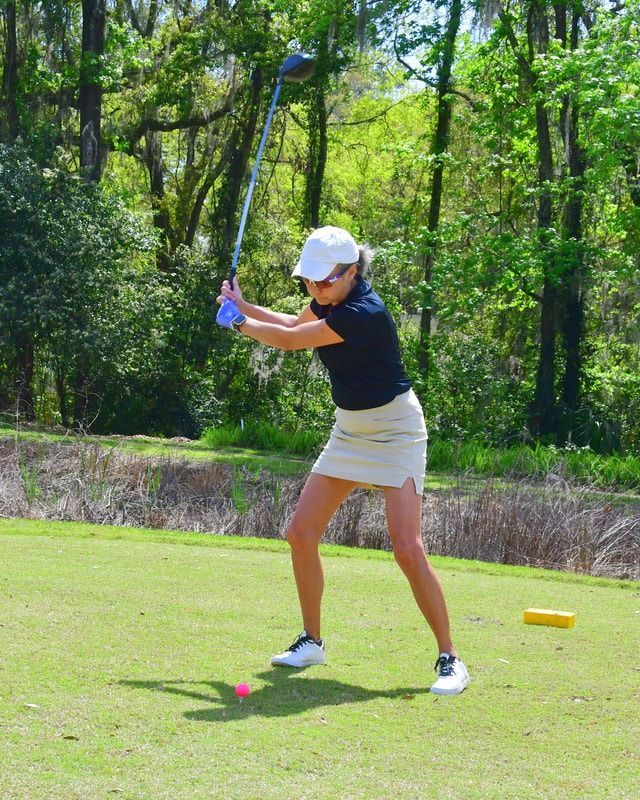Woman in golf attire swings a club on a green course, with trees in the background.