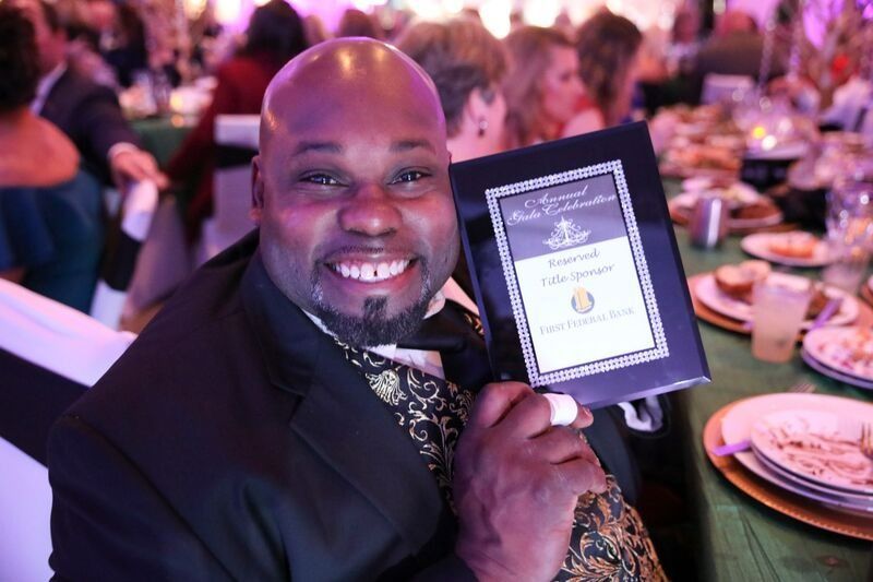 Man smiles, holding a framed award at a formal event, green and gold decor.