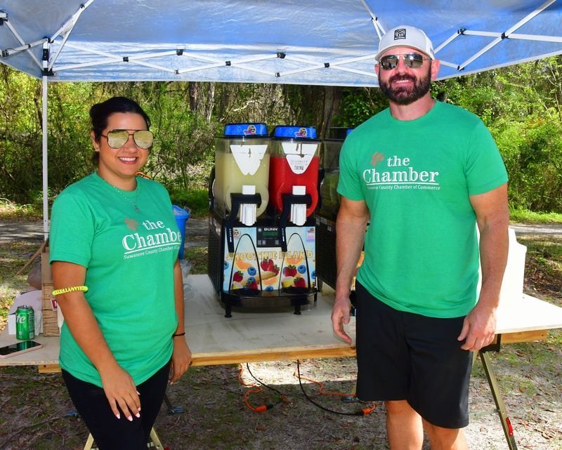 Two people in green shirts stand by a slushy machine outdoors.