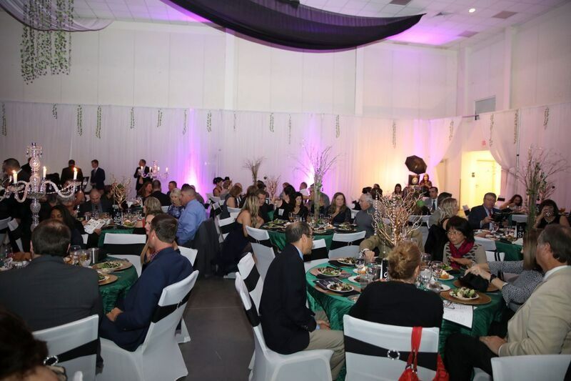 Guests seated at tables in a brightly lit banquet hall. Tables have green cloths and black chair sashes.