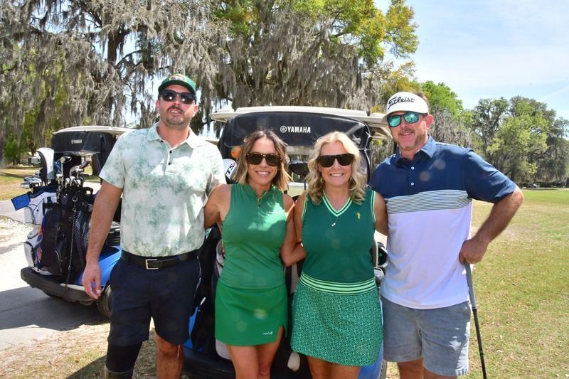 Four people in golf attire pose by golf carts on a sunny course.
