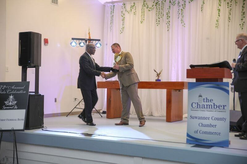 Two men shaking hands on stage at an event, likely an awards ceremony, with a Chamber of Commerce sign visible.