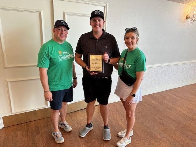 Three people pose indoors: one holds an award. Two wear green shirts, the other wears a black shirt and hat.