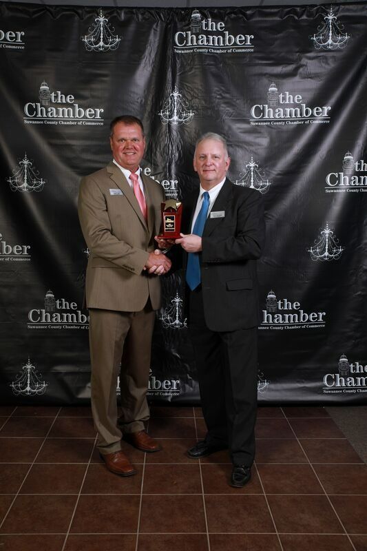 Two men in suits shaking hands, holding an award. Black backdrop with 