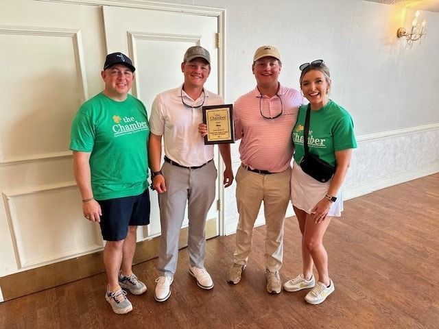 Four people indoors holding an award. Two wear green shirts. Two others wear golf attire. All are smiling.