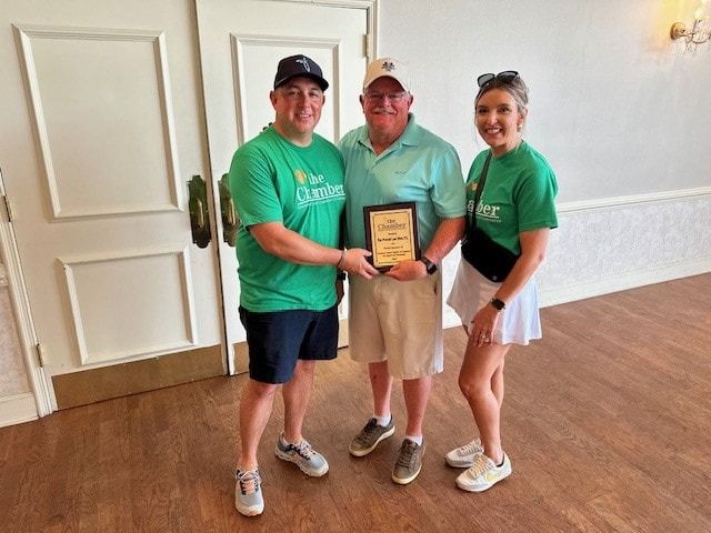 Three people holding plaque, wearing green shirts. Man in center smiles, receiving award. Light-colored room.